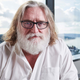 A photo of Gabe Newell with long hair, a big white beard and black glasses.